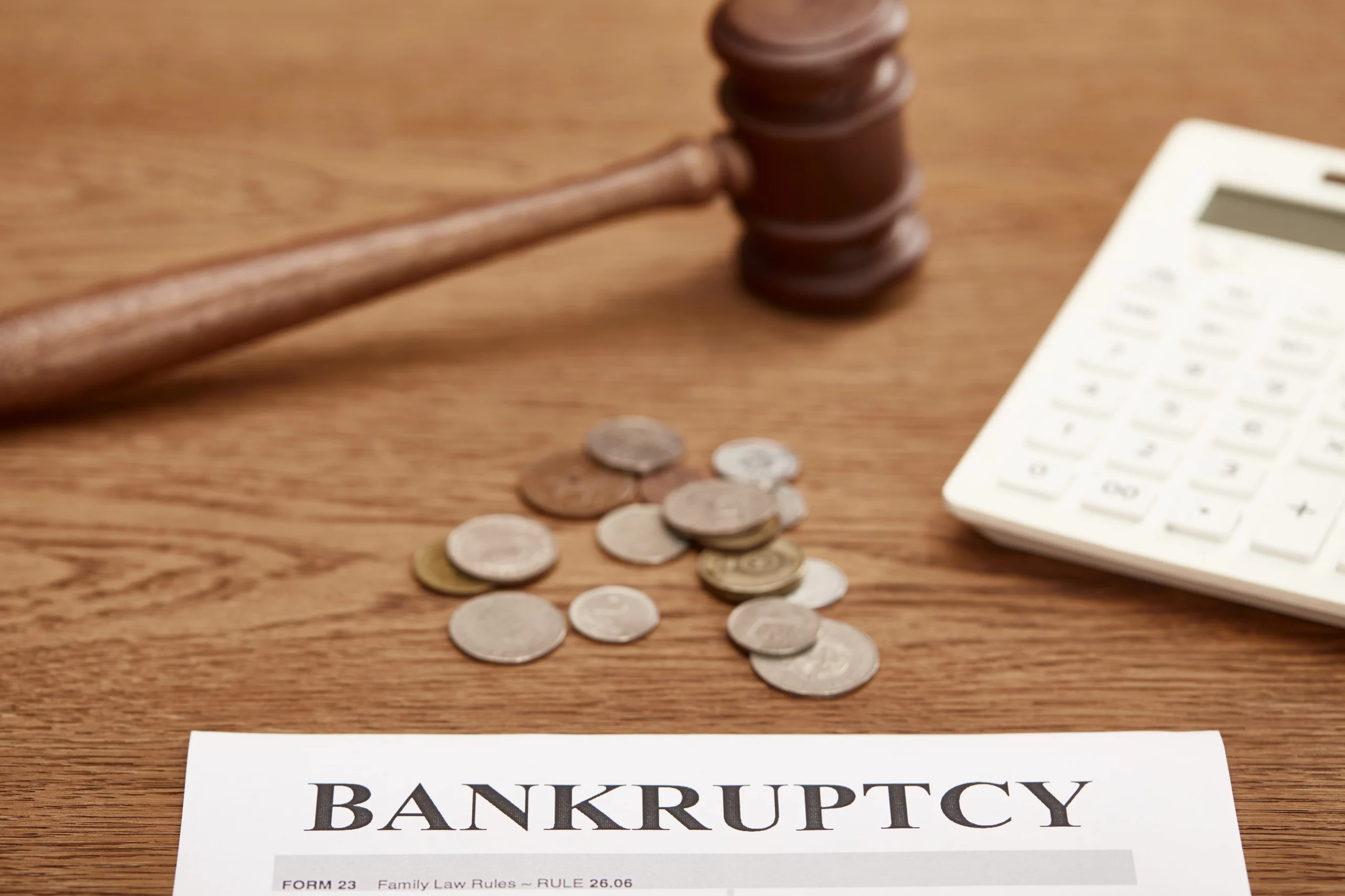 A legal document with "BANKRUPTCY" prominently printed is on a wooden desk. A wooden gavel, a pile of assorted coins, and a white calculator are also on the desk. The image symbolizes the legal and financial aspects of bankruptcy, representing the formal declaration and potential financial implications.