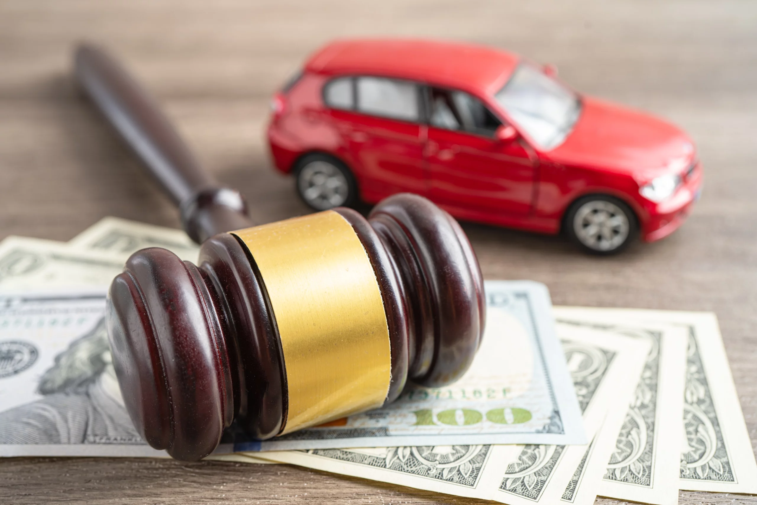 A wooden gavel rests on a scattering of US dollar bills, with a blurred red toy car in the background, all on a wooden surface. The image symbolizes legal and financial issues related to vehicle ownership, potentially representing car repossession, legal disputes over car loans, or insurance claims.