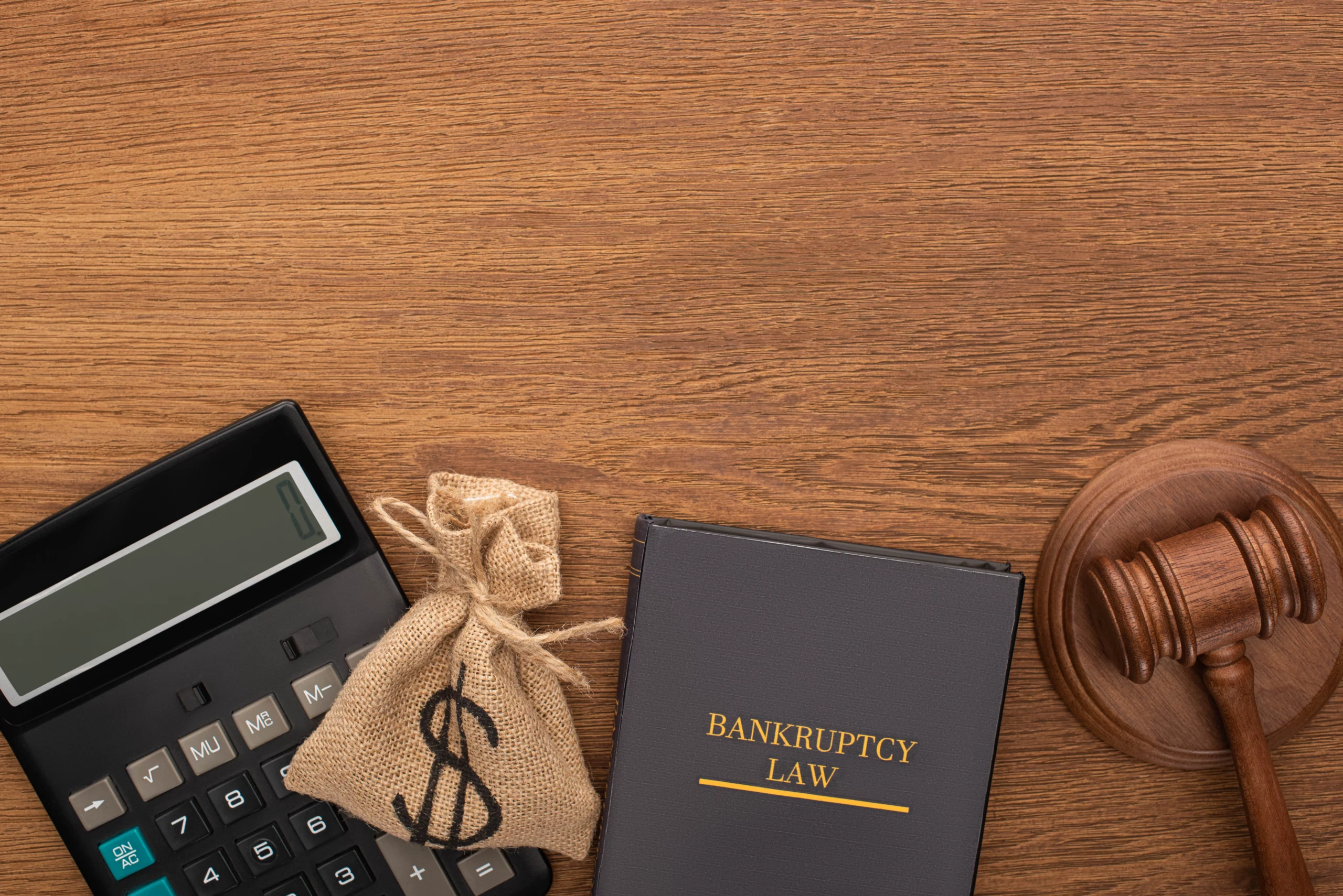 A top-down view of a wooden table shows a black calculator, a small burlap money bag with a dollar sign, a book titled "BANKRUPTCY LAW," and a wooden gavel. These objects are arranged in the bottom half of the image, leaving ample space above. The image symbolizes the legal and financial tools associated with bankruptcy, legal advice, and debt management.