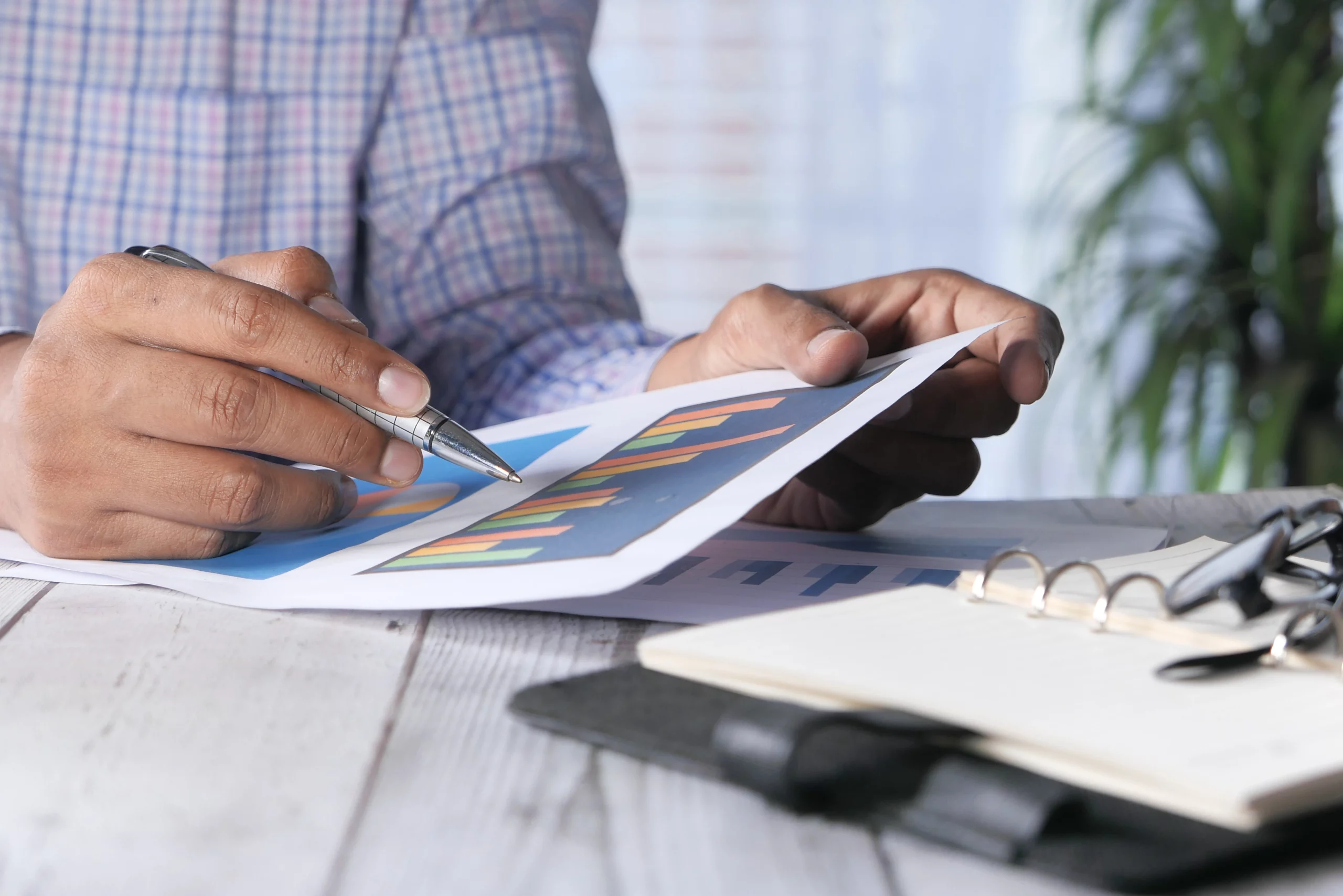 Person in a plaid shirt reviewing bar charts with a pen, an open notebook, and glasses on a wooden table, symbolizing financial planning.