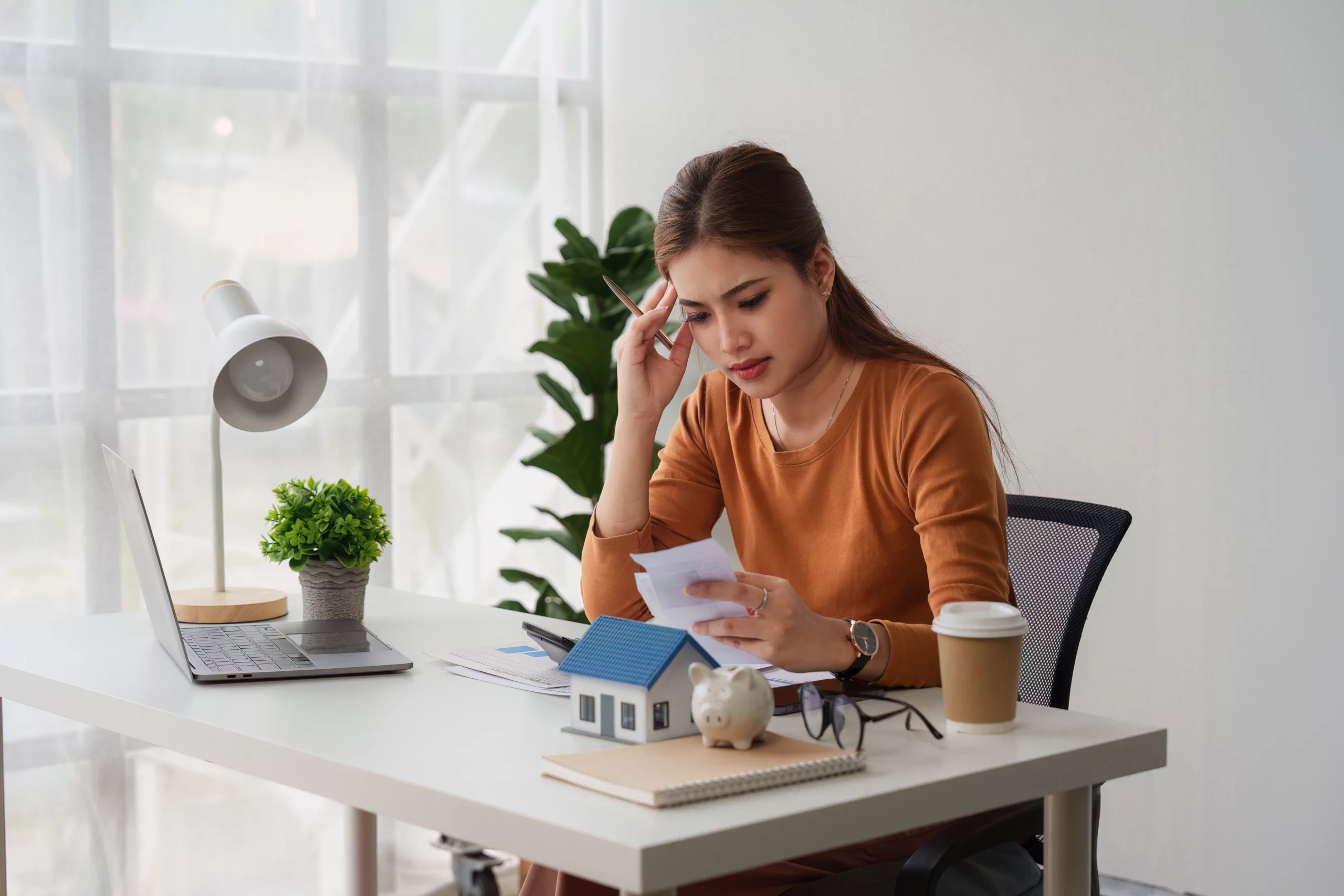 Woman in a brown sweater reading documents at a desk with a laptop, house model, piggy bank, and coffee cup in a bright room.