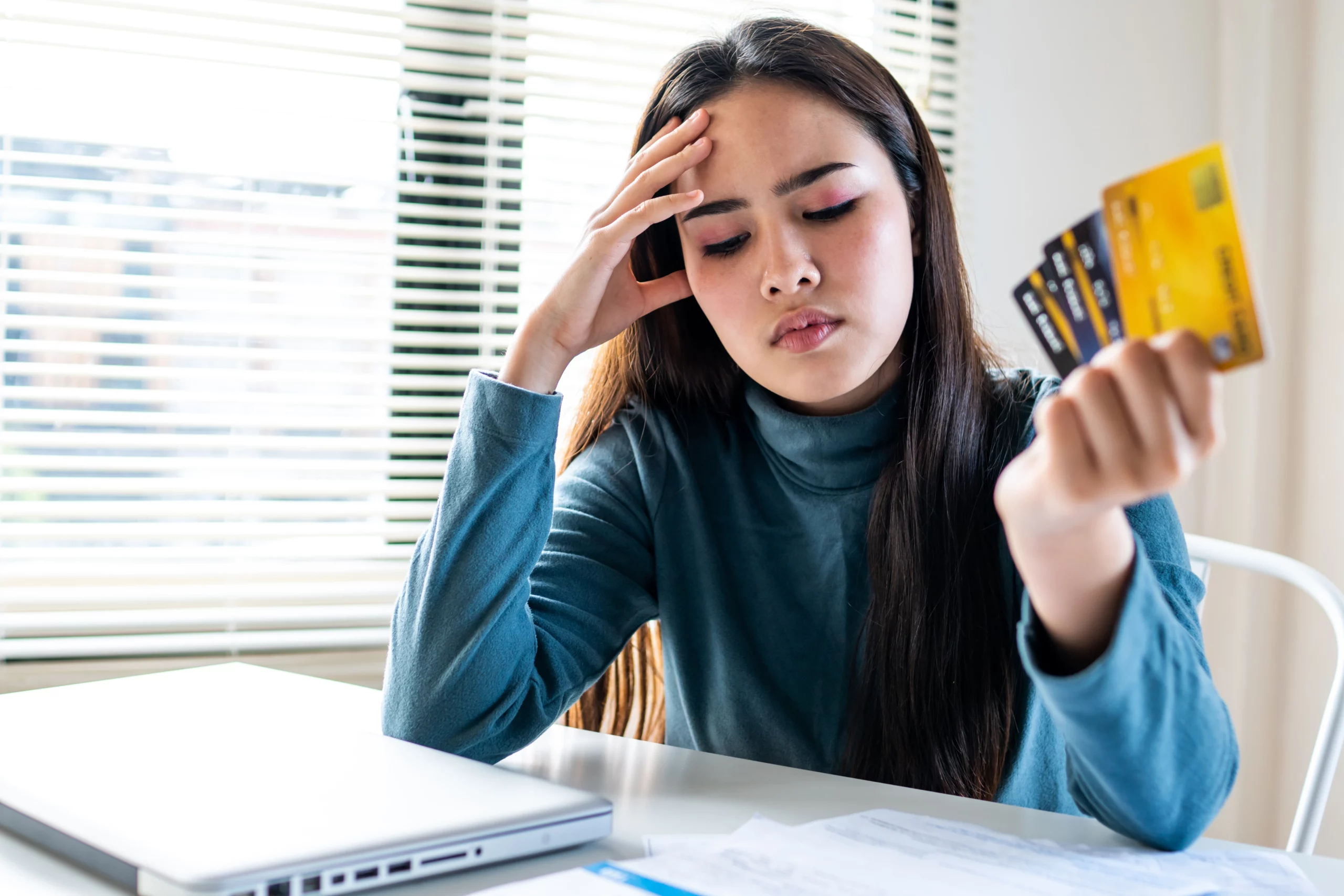 Young woman in blue sweater holding multiple credit cards, stressed, sitting at table with laptop and bills in Oregon