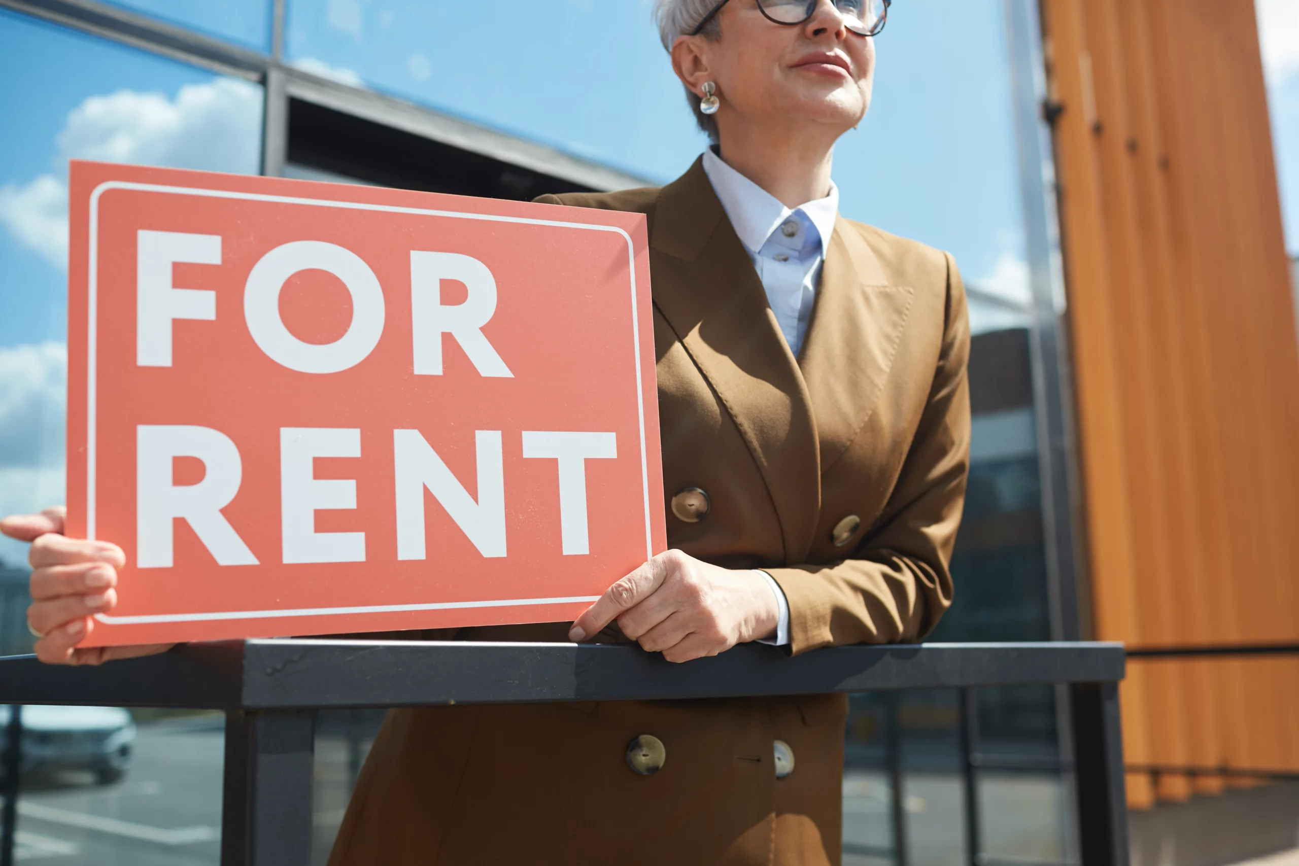 Confident landlord in brown blazer holding a red "For Rent" sign in front of a modern Portland building under a clear blue sky.