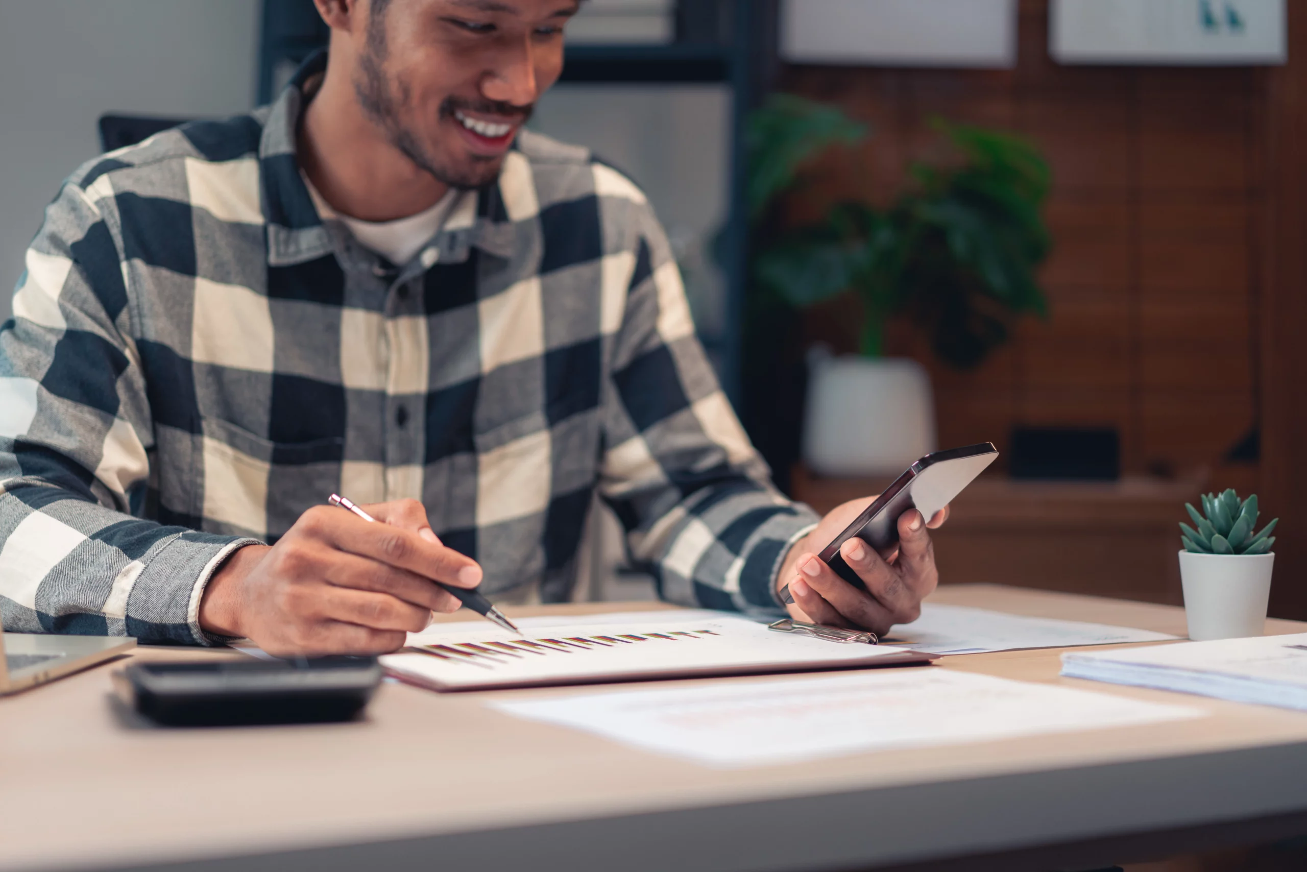 Smiling person in a plaid shirt using a smartphone and pointing at financial graphs on a clipboard, with a calculator and plant on a desk in a cozy home office.