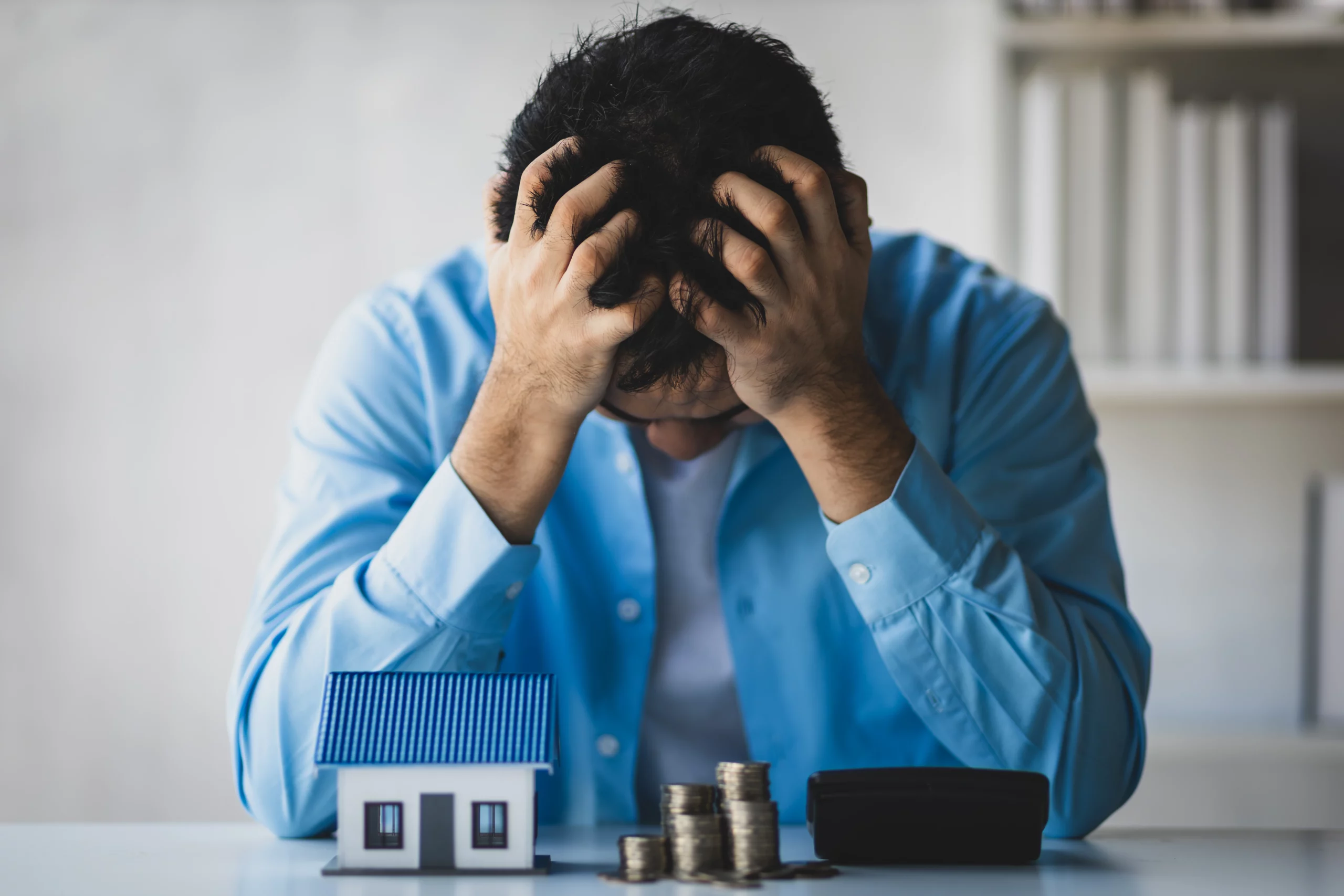 Man in blue shirt with head in hands, sitting at a table with a small house model and stacked coins, conveying financial stress about keeping a home during Chapter 7 bankruptcy in Oregon.