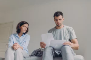 Man and woman sitting on a bed in a neutral-toned room, looking concerned — the man reading papers while the woman watches him anxiously, suggesting financial stress and uncertainty about filing Chapter 13 bankruptcy without a spouse.